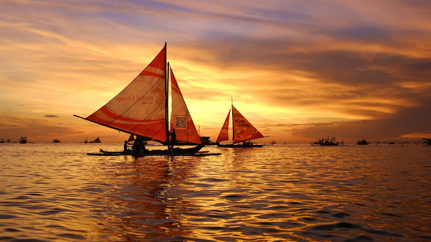 Traditional paraw sailboats with red sails on calm water off Boracay at sunset, the sky burning orange behind them.