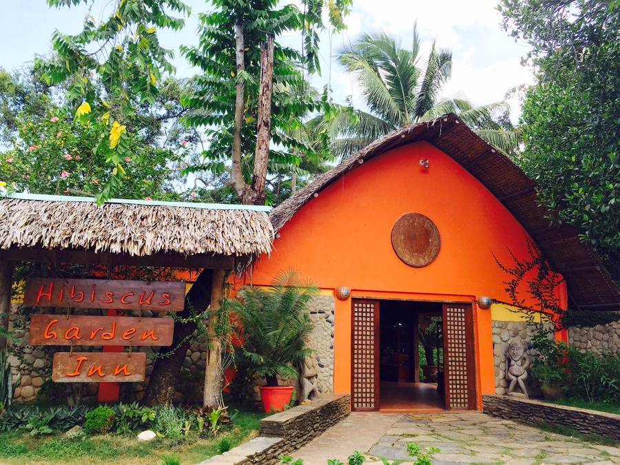 The entrance to Hibiscus Garden in Puerto Princesa: an orange building under a thatched roof behind a carved wooden sign, set in tropical planting.