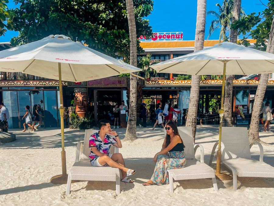 Red Coconut Beach Hotel from White Beach: guests on loungers under branded umbrellas, the hotel's red sign above its beachfront restaurant.